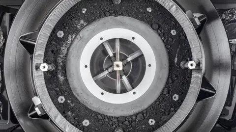 NASA/Erika Blumenfeld/Joseph Aebersold Nasa capsule that looks like an alloy wheel containing the black dusty sample of asteroid Bennu in a central section. The sample resembles coal dust with different sizes of fragments of black rock. There are also a number of bolts that were holding the capsule's lid in place.