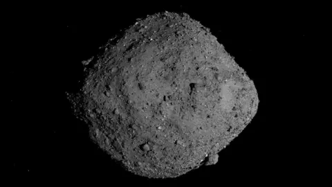 NASA/Goddard/University of Arizona Image of a 500m-wide asteroid called Bennu that looks like a grey rock which is wider in the middle than at each end. It isn't smooth -- there are different sized knobbly lumps sticking out from its surface.