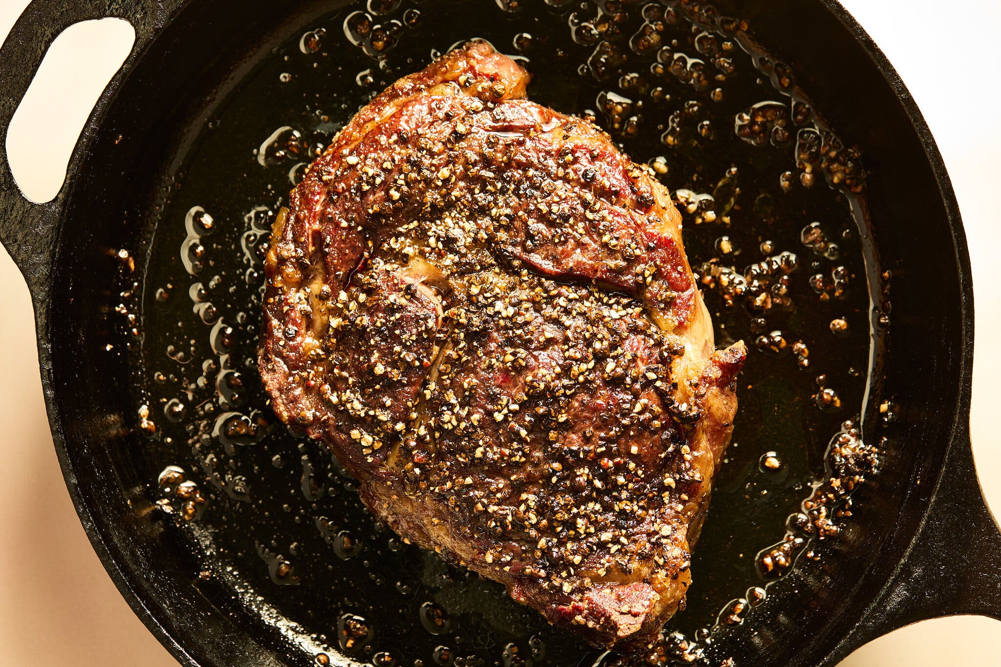 A steak coated in peppercorns cooks in a black cast iron skillet.