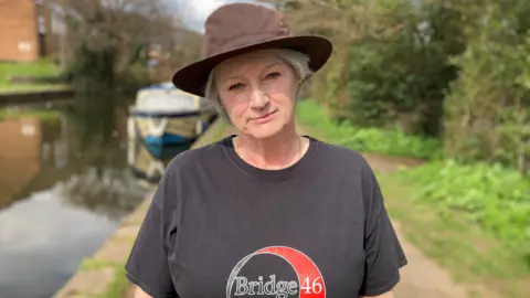 Tracey Leake-Jones of the Bridge 46 to Five Locks Canal Group looks straight at the camera. She is wearing a black T-shirt and a brown hat. She is standing in front of the canal and a blue canal boat at the Five Locks basin north of Cwmbran in Torfaen.