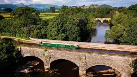 Beacon Park Boats / As You See It Media A green canal boat can be seen on an aqueduct where the Monmouthshire and Brecon Canal crosses the River Usk at Brynich near Brecon, Powys.