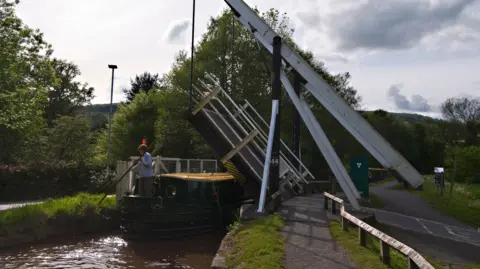 The Talybont drawbridge is raised for holiday hire barges on the Monmouthshire and Brecon Canal at Talybont, Powys. A black canalboat with a yellow roof is passing under the raised bridge, with a person wearing a striped top and holding a large pole standing at the front of a boat.