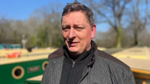 Alasdair Kirkpatrick, owner of Beacon Park Boats stands on a barge wearing a grey coat and jacket. In the background you can see other green barges parked up.