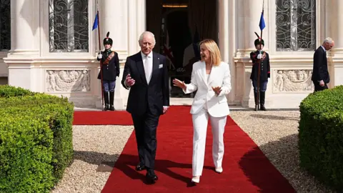 PA Media King Charles, in a black suit and white shirt with a silver tie, walks with Giorgia Meloni, who wears a white suit, at Villa Doria Pamphili, in Rome,