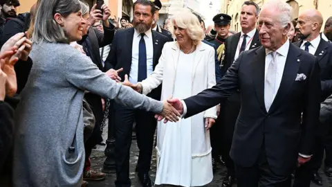 Getty Images The Queen and King shake hands with people in the crowd. The King is wearing a dark-coloured suit with a white shirt and the Queen is wearing a long white dress with a white coat