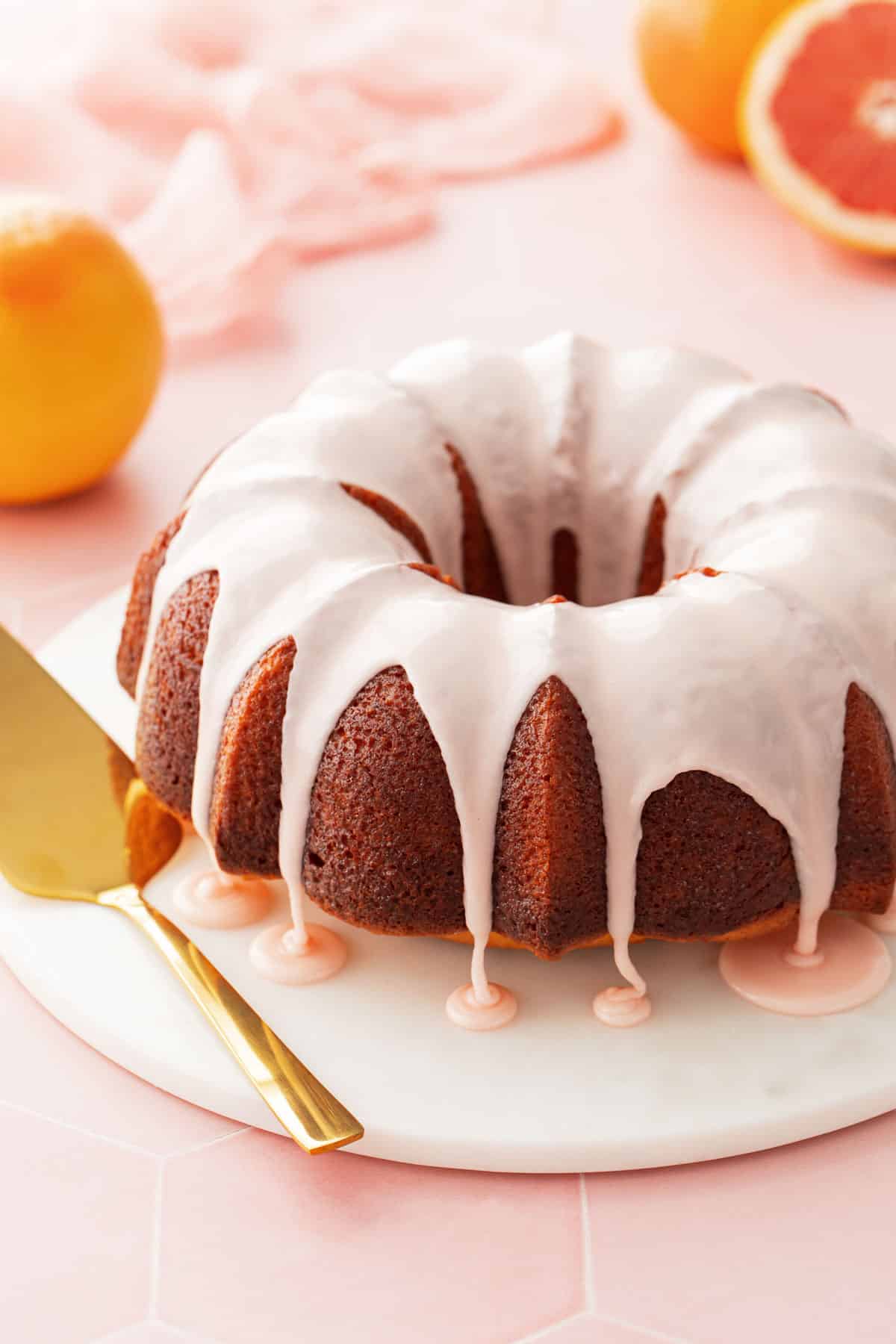 Pink glazed grapefruit bundt cake on a marble cake plate with a pink background and a cut pink grapefruit in the background.