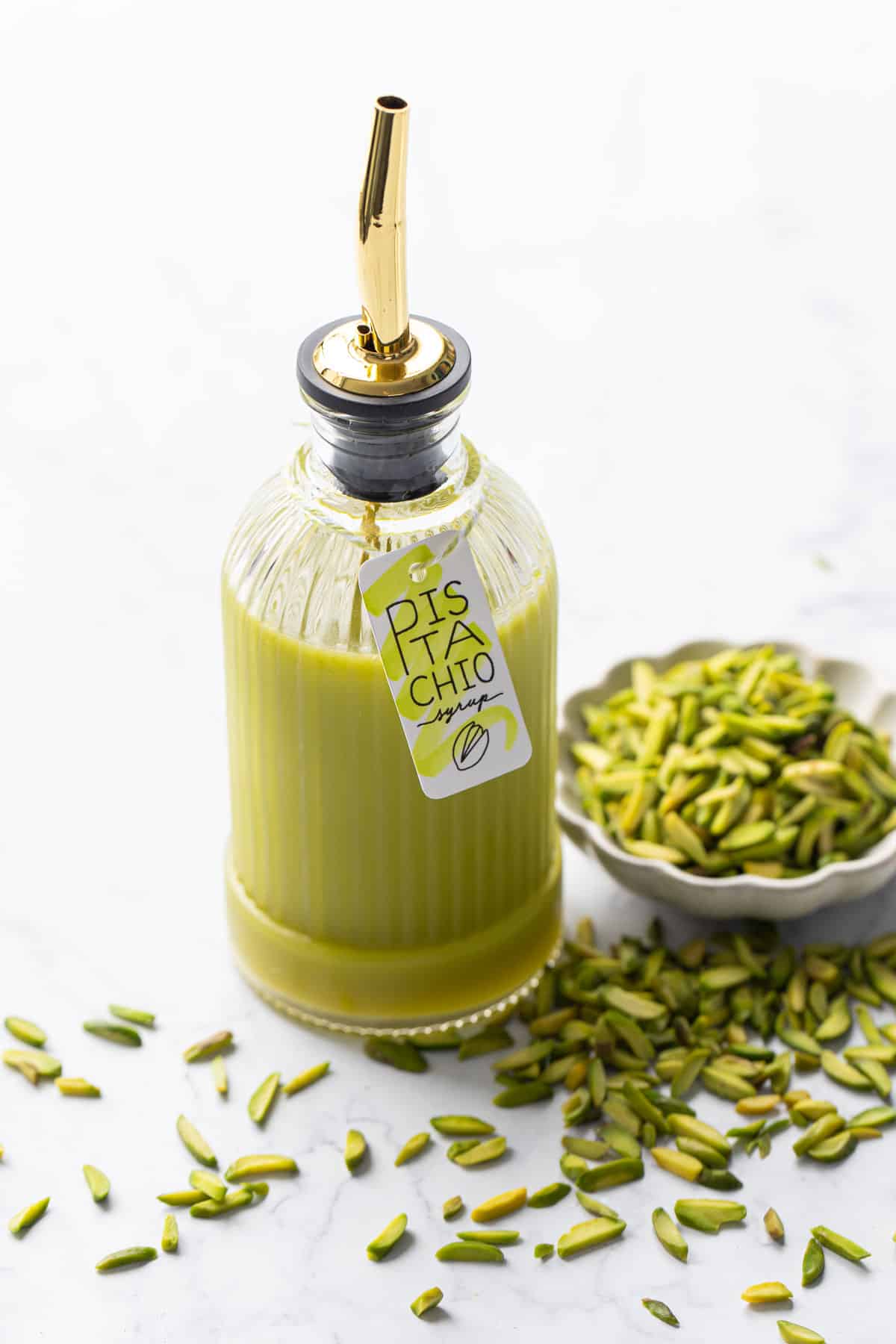 Glass bottle with green pistachio syrup and a cute tag tied on with twine, with a bowl of slivered pistachios on the side.