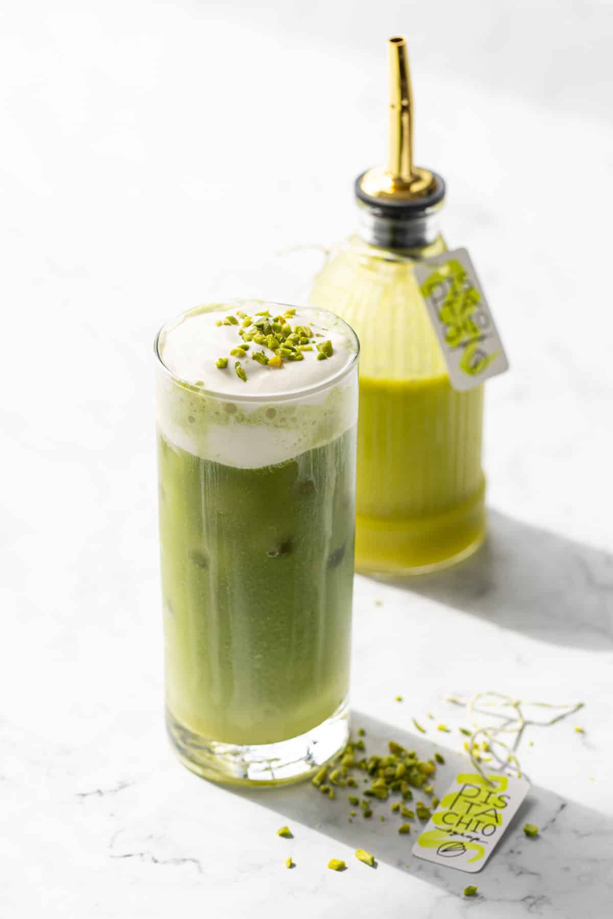 Pistachio matcha iced latte in a glass with a bottle of homemade pistachio syrup in the background.