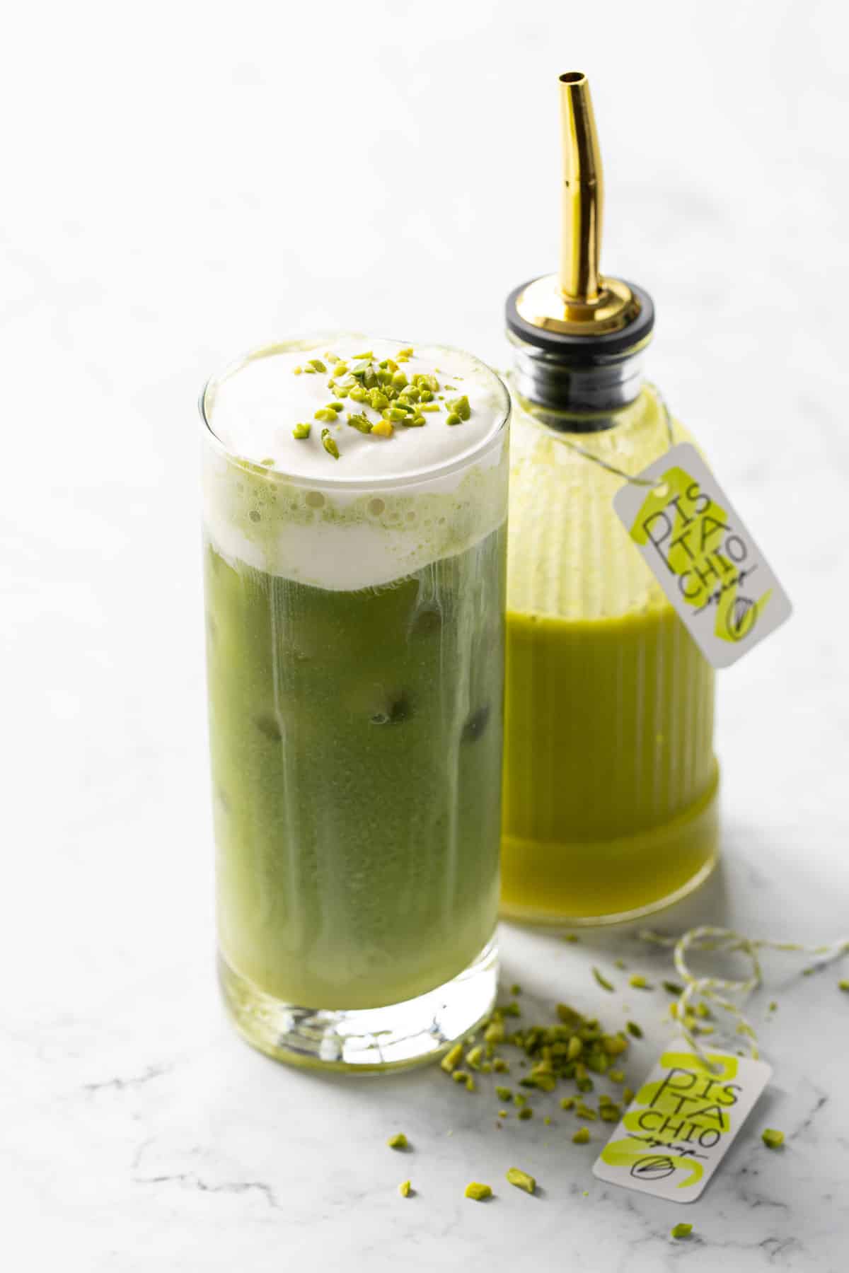 Pistachio matcha latte topped with pistachio cold foam, with a faceted glass bottle of pistachio syrup in the background.