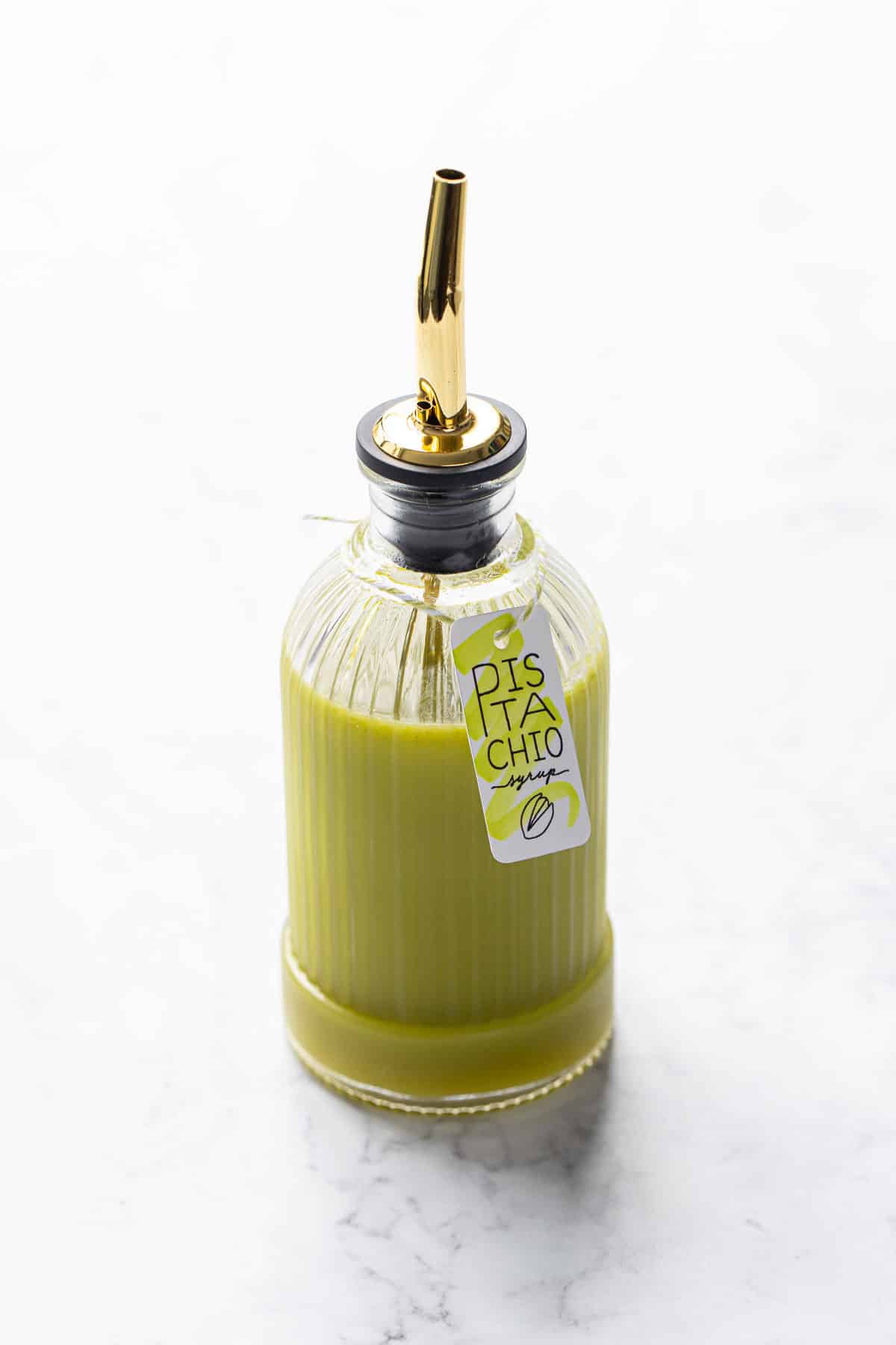 Faceted glass bottle filled with Homemade Pistachio Syrup on a white marble background, with a printed tag tied on the neck with twine.