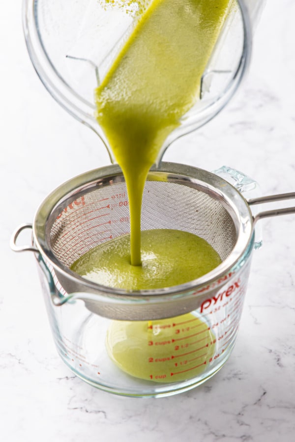Pouring blended pistachio syrup through a fine mesh sieve into a spouted measuring cup.
