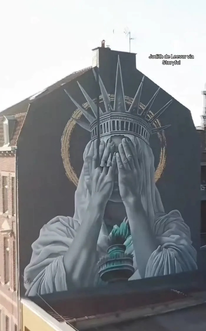 Mural of the Statue of Liberty covering its eyes, painted on a building. Artist's name is visible in the upper right corner