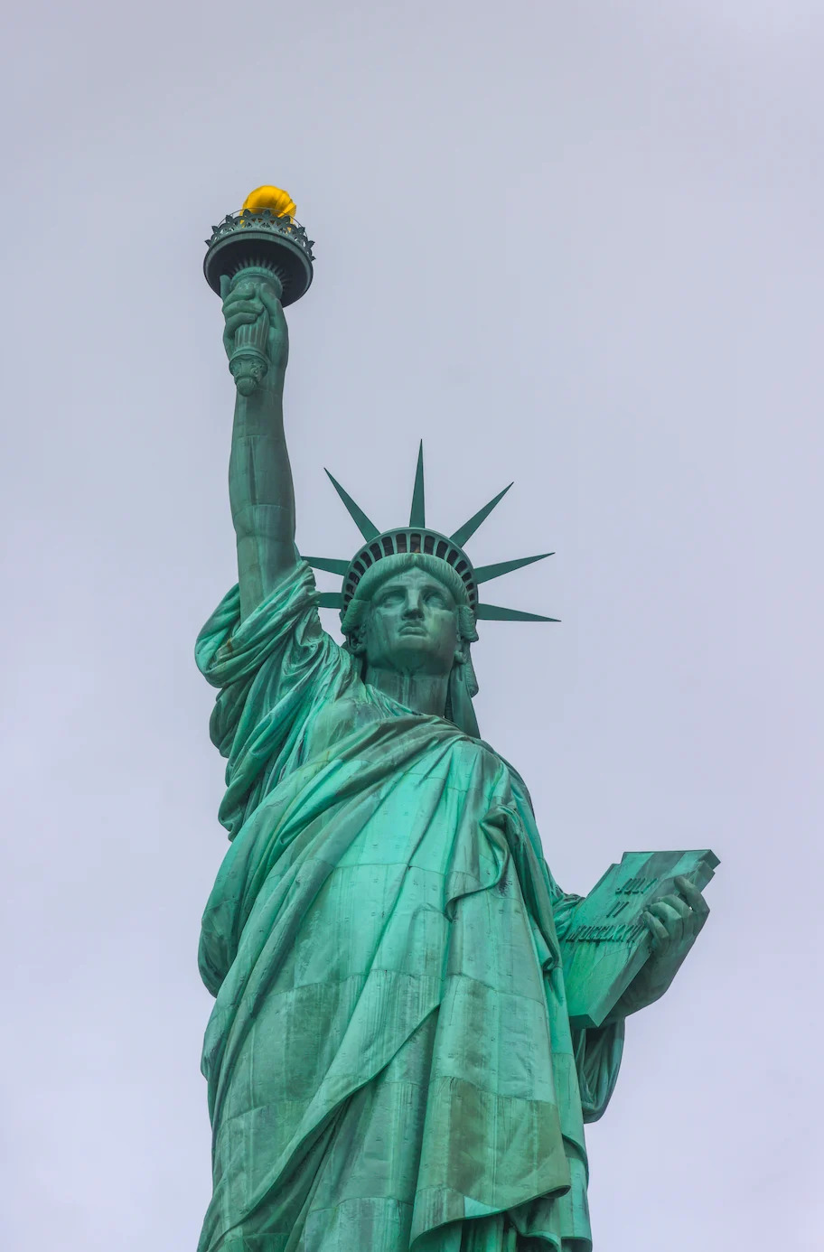 The Statue of Liberty holding a torch and a tablet, viewed from below, set against a cloudy sky