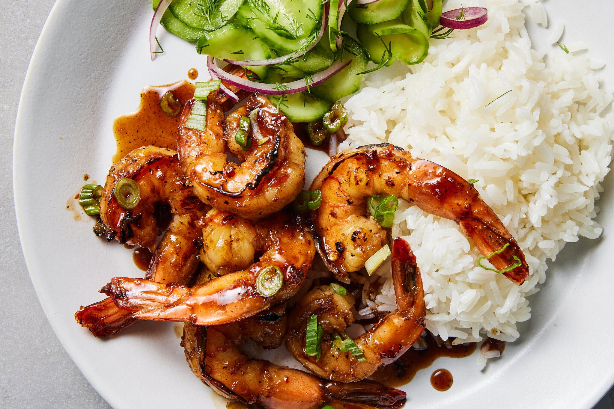 An overhead image of sauced tail-on shrimp on a bed of rice next to sliced cucumbers and red onion.