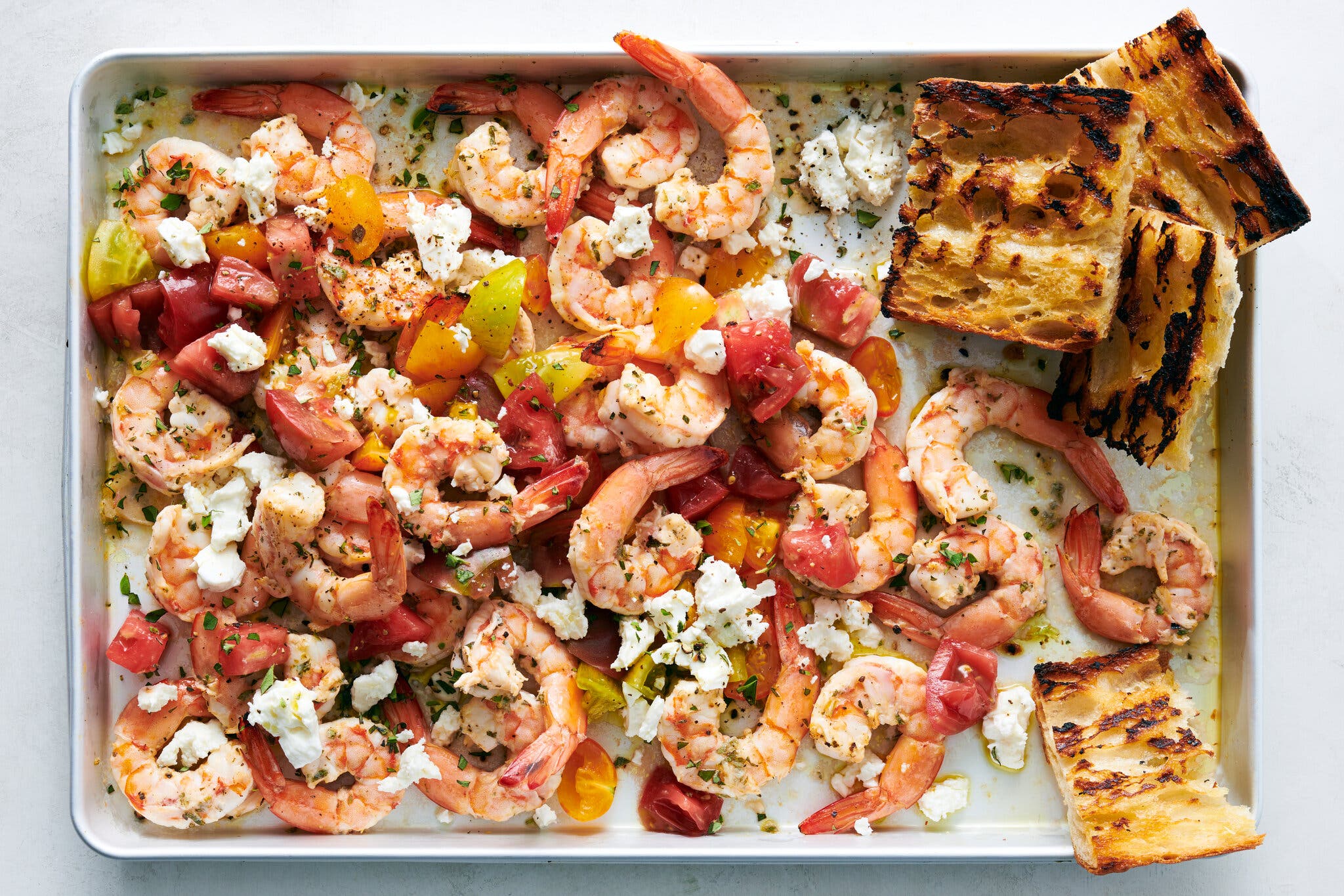 An overhead image of a rimmed sheet pan filled with shrimp, tomatoes and feta. A few pieces of bread sit off to one corner.