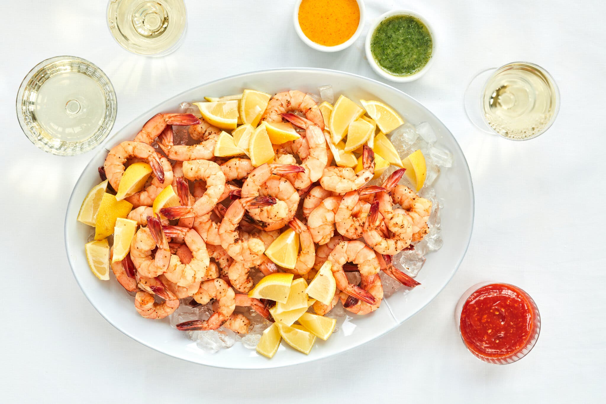 A white oval platter is piled high with poached shrimp and lemon wedges over ice. It’s surrounded by dipping sauce and white wine.