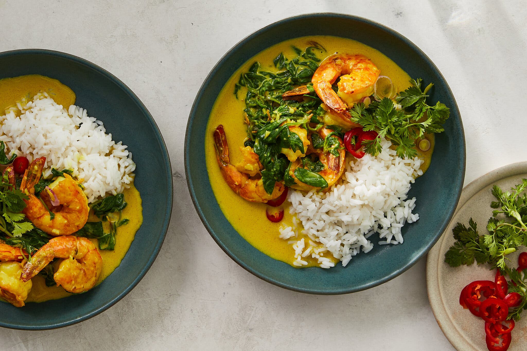 Two slate-blue bowls are filled with golden pools of sauce and shrimp, greens, and rice.