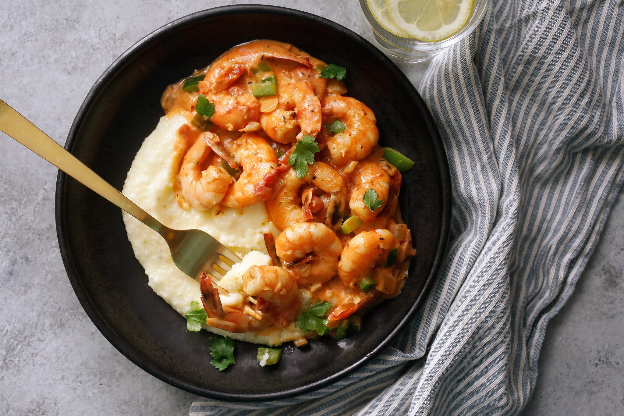 Saucy shrimp are set over grits in a black bowl with a golden spoon.