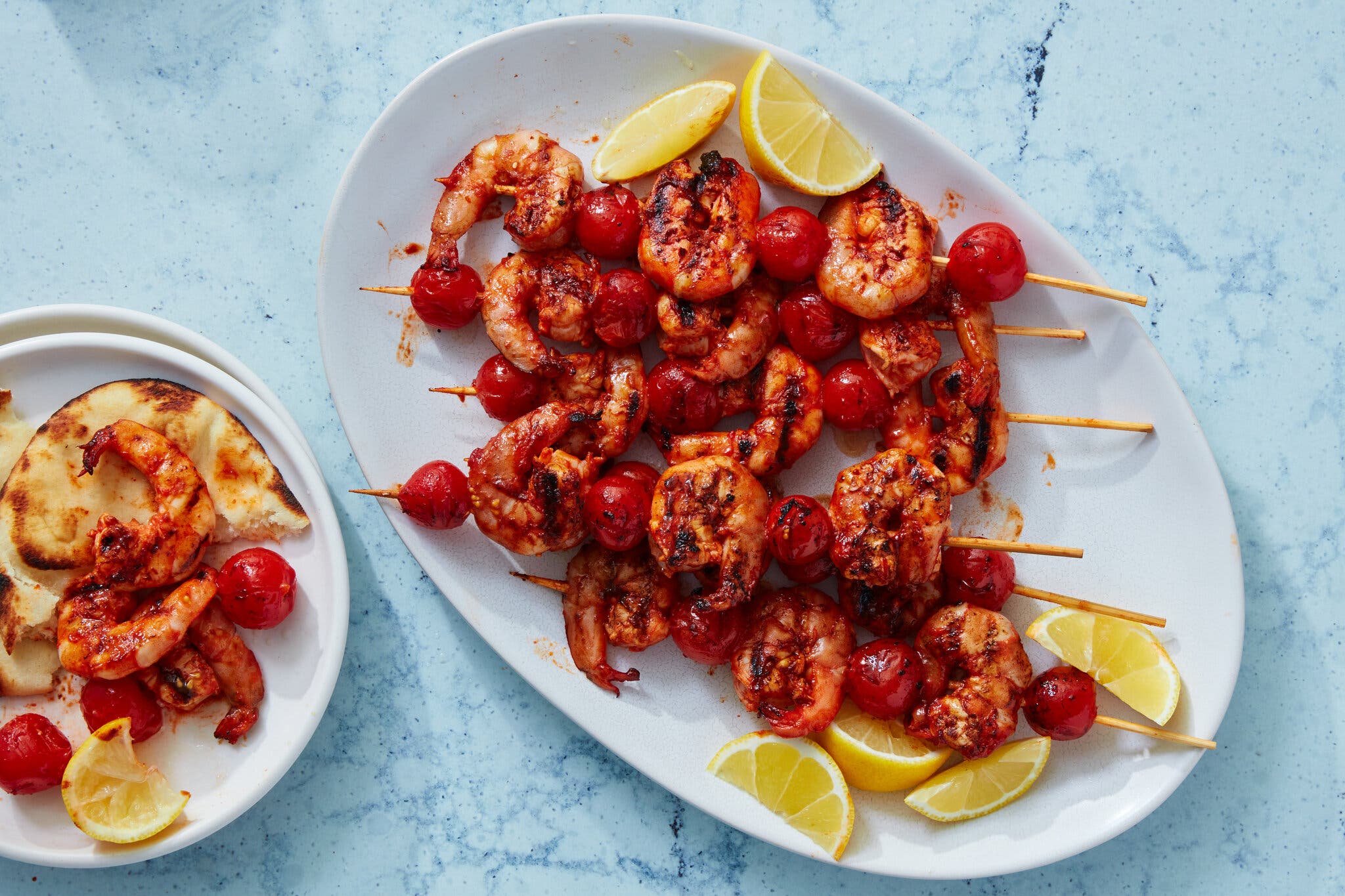 A white oval platter holds lemon wedges and skewers of sauced griilled shrimp and cherry tomatoes.