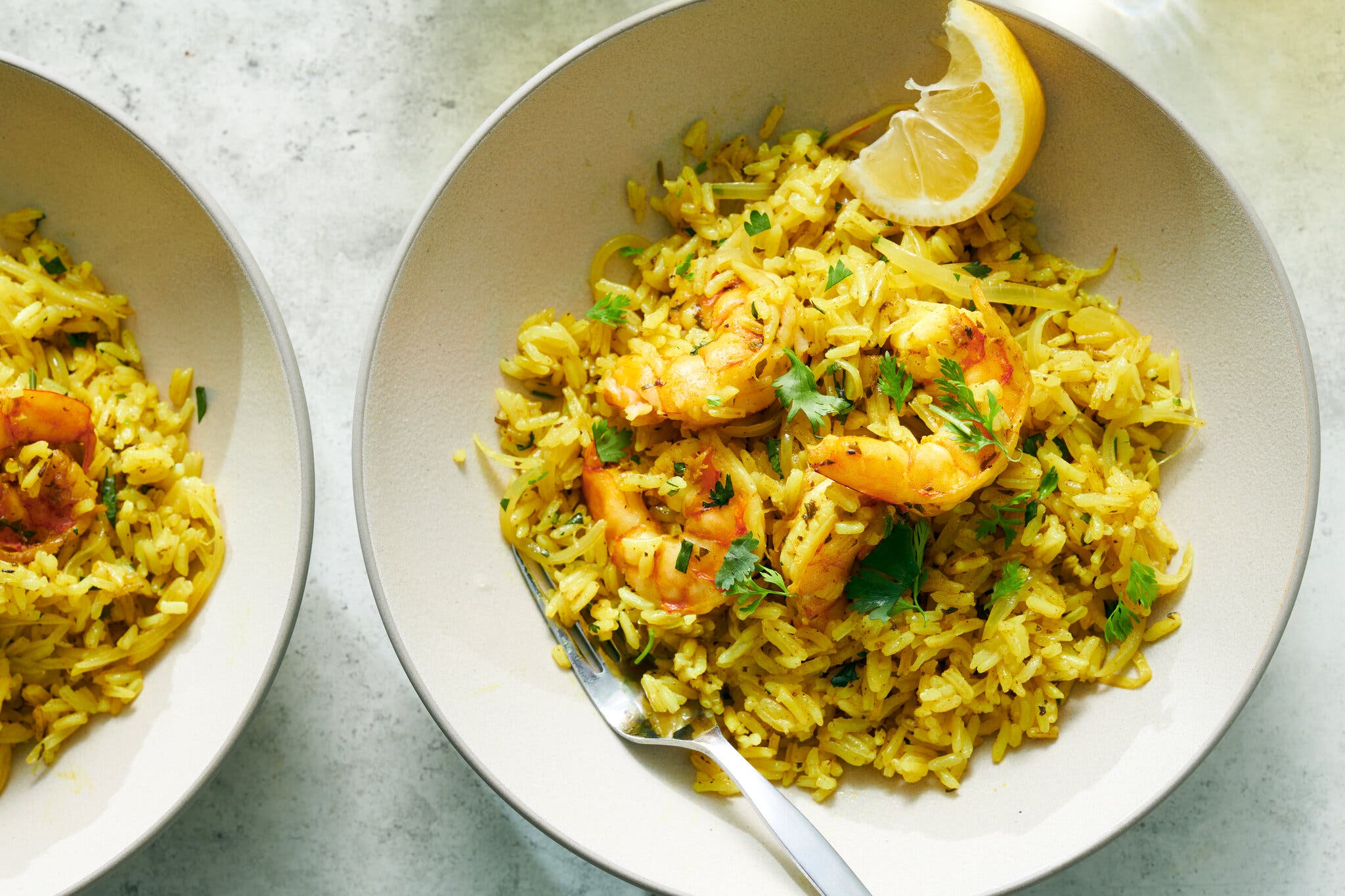 A mound of golden rice and shrimp fills a white bowl that also holds a fork and lemon wedge.