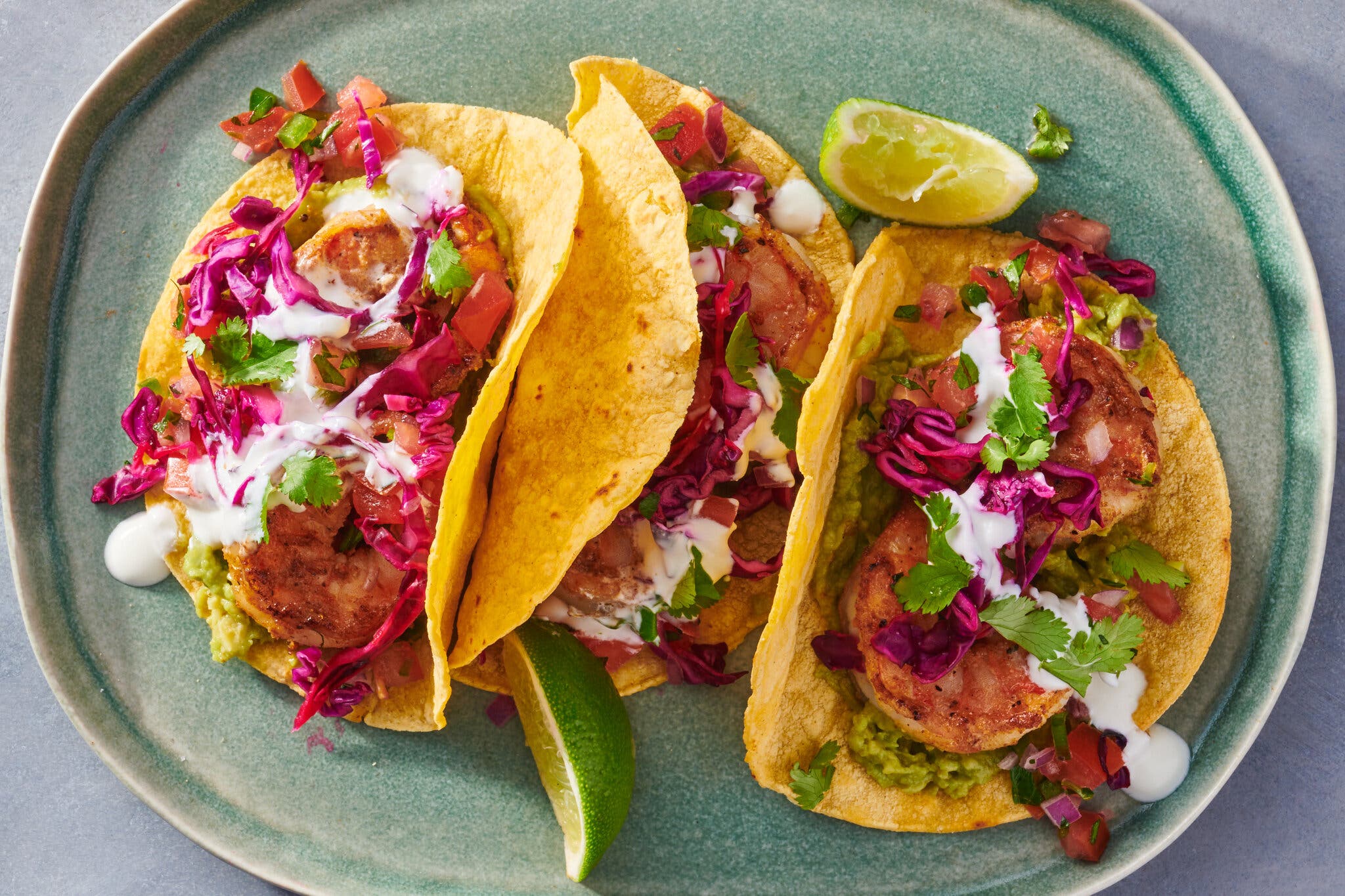 Three tacos filled with shrimp, white sauce, red cabbage, guacamole and cilantro are lined up on a blue-green plate.