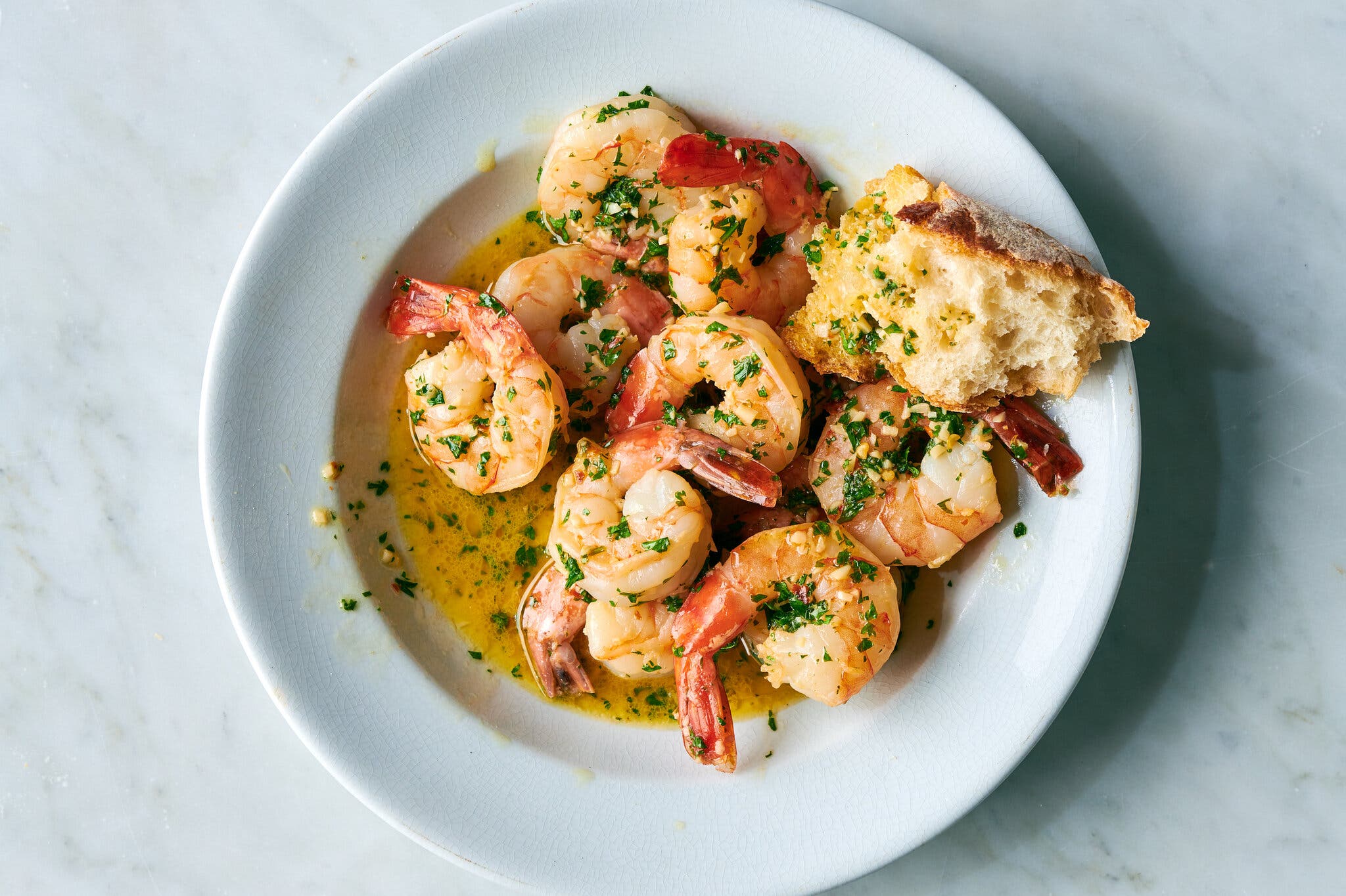 A white dinner plate is filled with parsley- and garlic-flecked shrimp and a hunk of baguette.