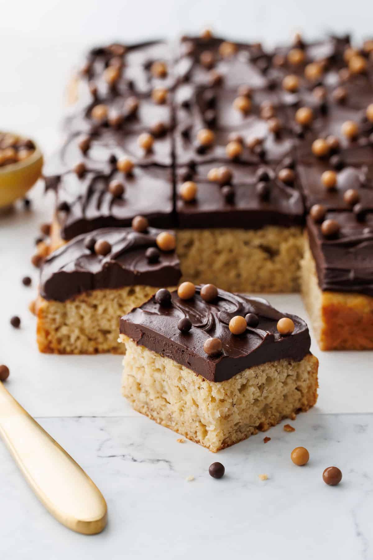 Cut piece of Brown Butter Banana Cake with Chocolate Fudge Frosting on a piece of parchment with a gold knife and bowl of crisp pearls in the background.
