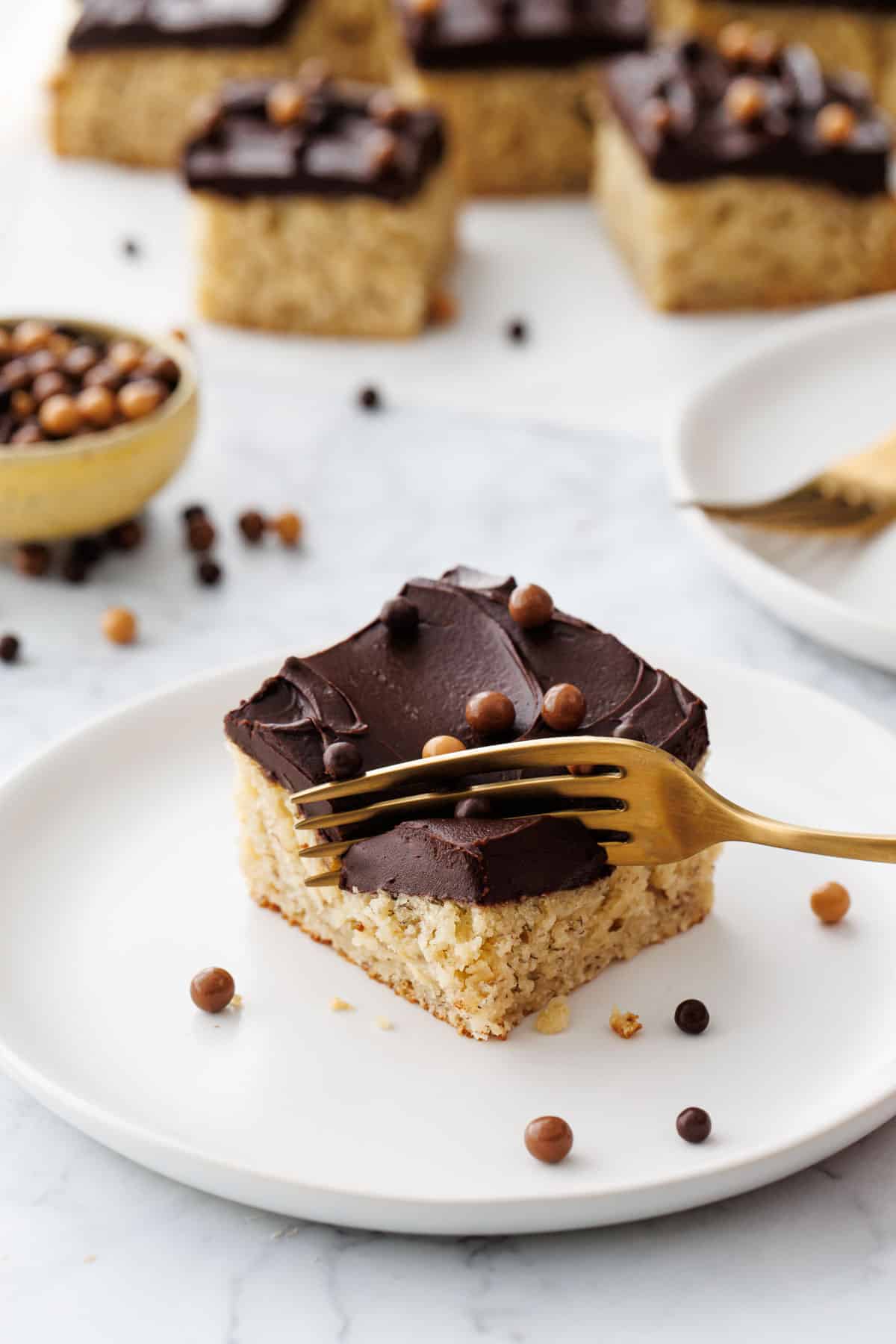 Gold fork cutting a corner of Brown Butter Banana Cake with Chocolate Fudge Frosting to show the tender texture and luscious chocolate frosting.