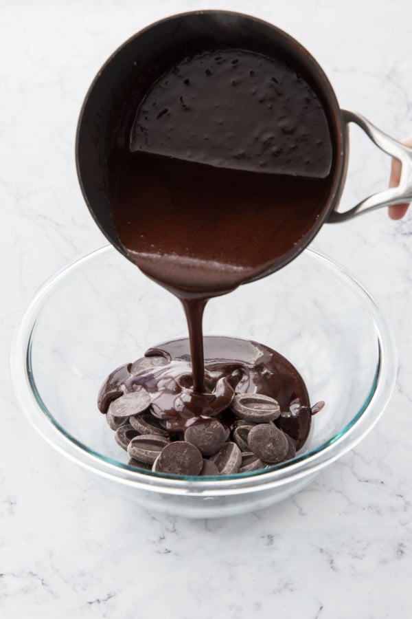 Pouring hot cream-cocoa mixture over chopped chocolate in a glass bowl.