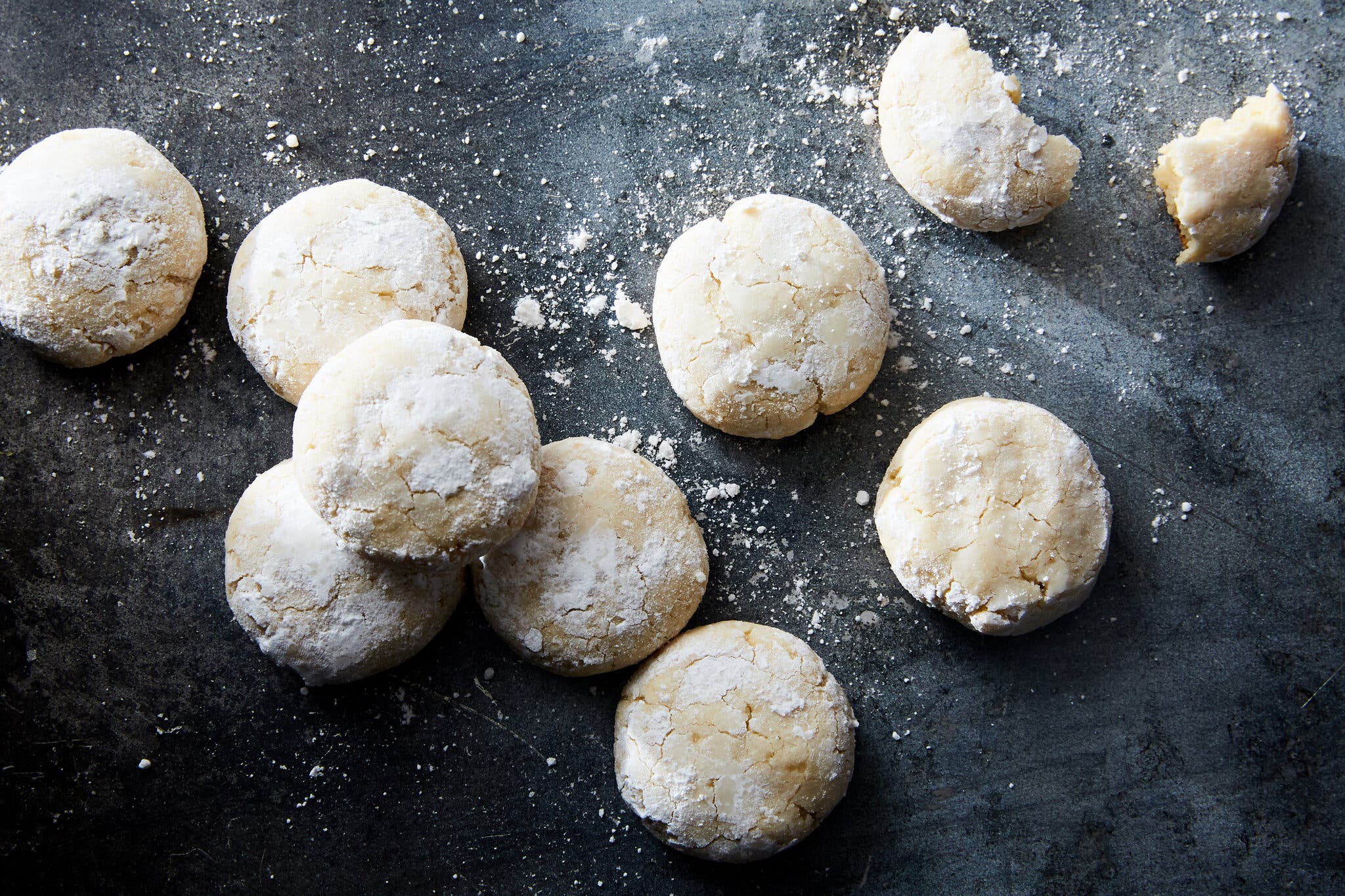 A close-up of several powdered sugar cookies on a dark, textured surface, with one cookie broken.