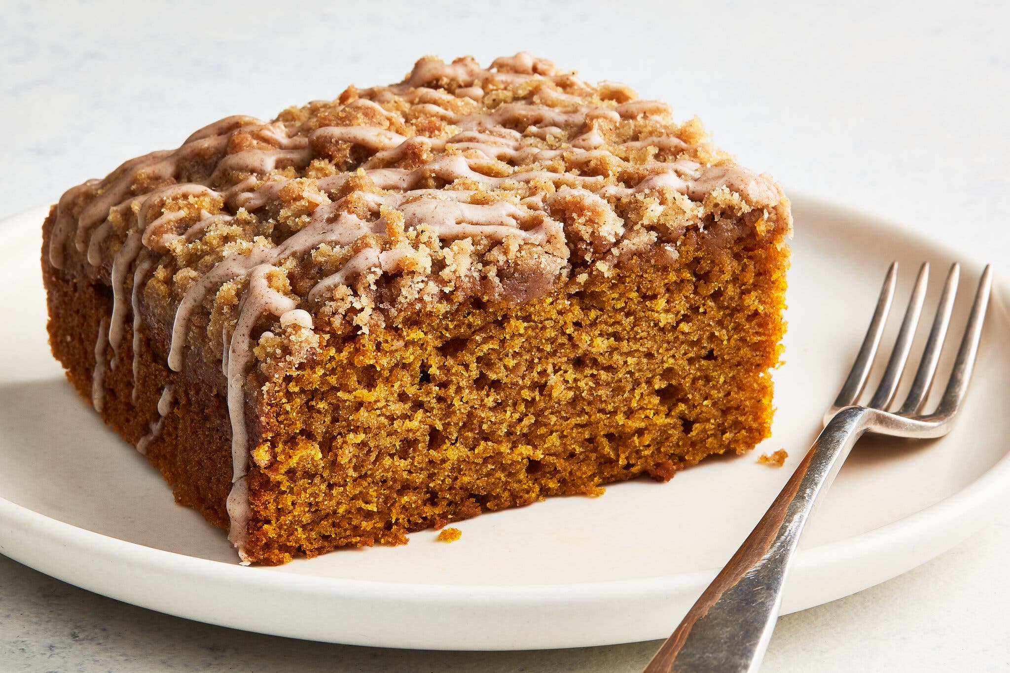 A slice of pumpkin coffee cake with a streusel topping and white icing drizzle on a cream plate next to a fork.