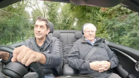 BBC climate editor Justin Rowlatt, dressed in black puffer coat, drives an open-top car down a country lane, beside economist Paul Cheshire in the passenger seat, whose hands are folded across his stomach