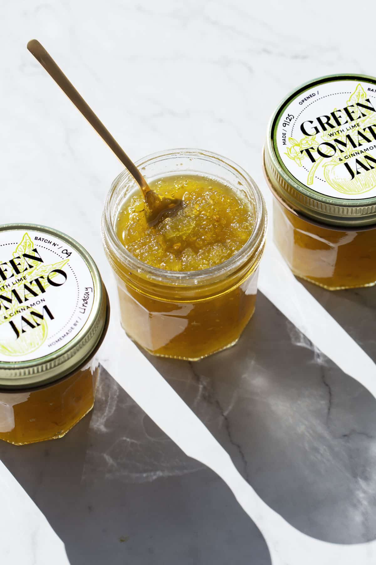 Three jars of Green Tomato Jam with designed printable labels, one jar open with a spoon in it to show the final jammy texture.