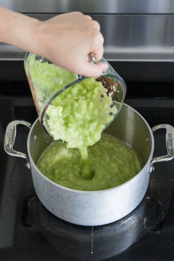 Pouring green tomato puree into a saucepan on the stovetop.