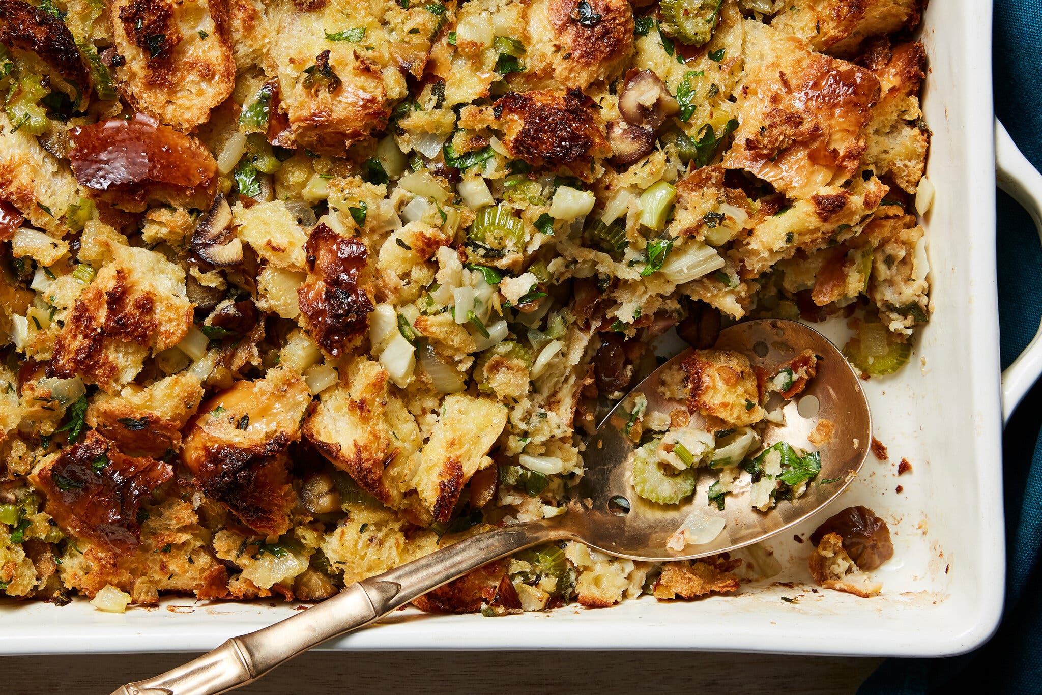 Golden-brown stuffing with chunks of bread, green celery, onions and mushrooms fills a white baking dish, with a spoon in one corner.