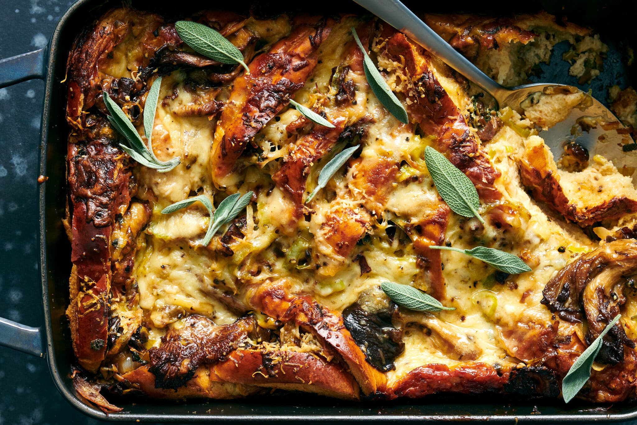 A golden-brown baked bread casserole in a dark pan, topped with melted cheese and fresh sage.