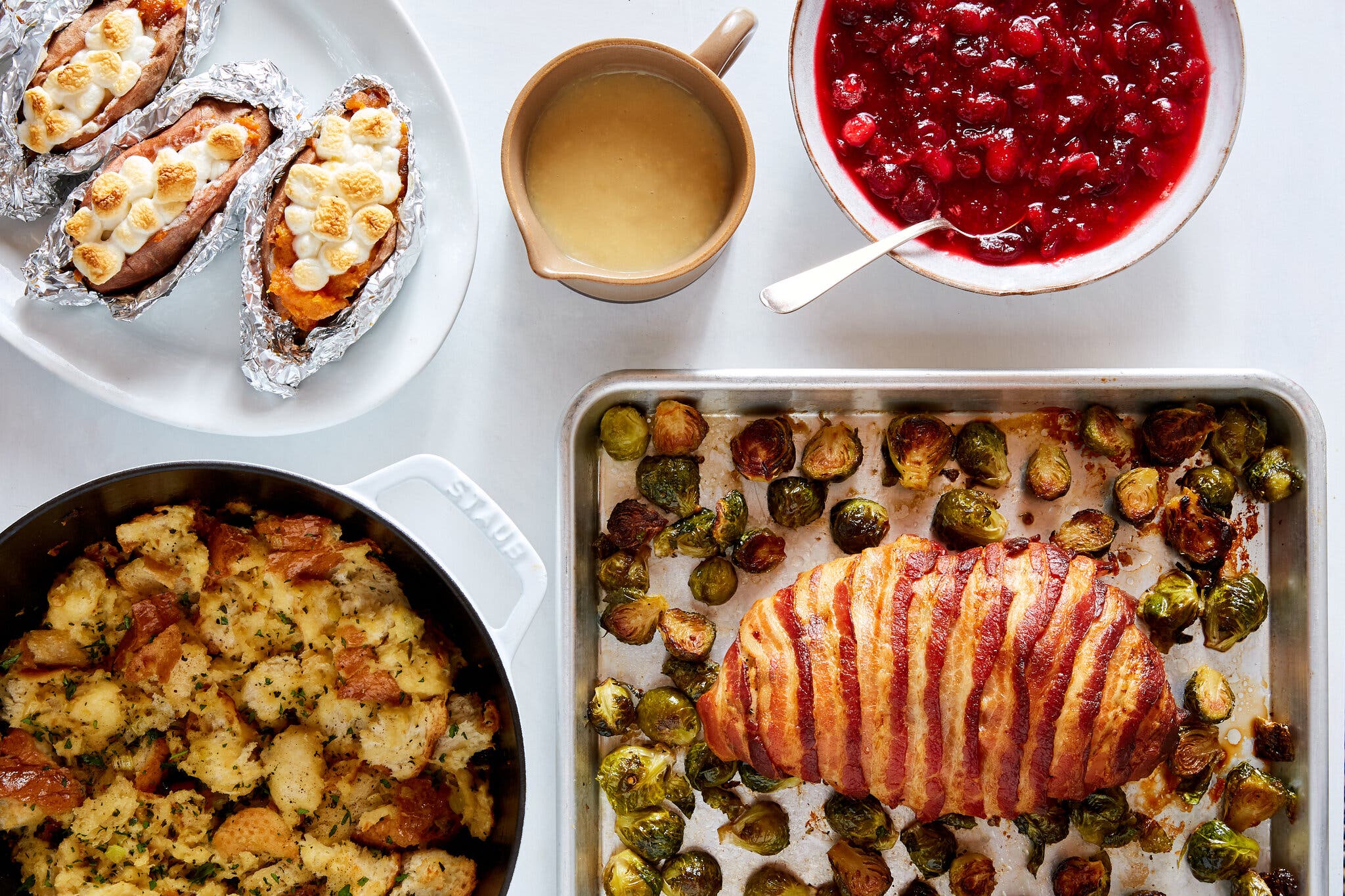 A spread of food on a white table including a bacon-wrapped turkey breast on a tray with brussels sprouts, sweet potatoes with marshmallows, red cranberry sauce, gravy and stuffing.