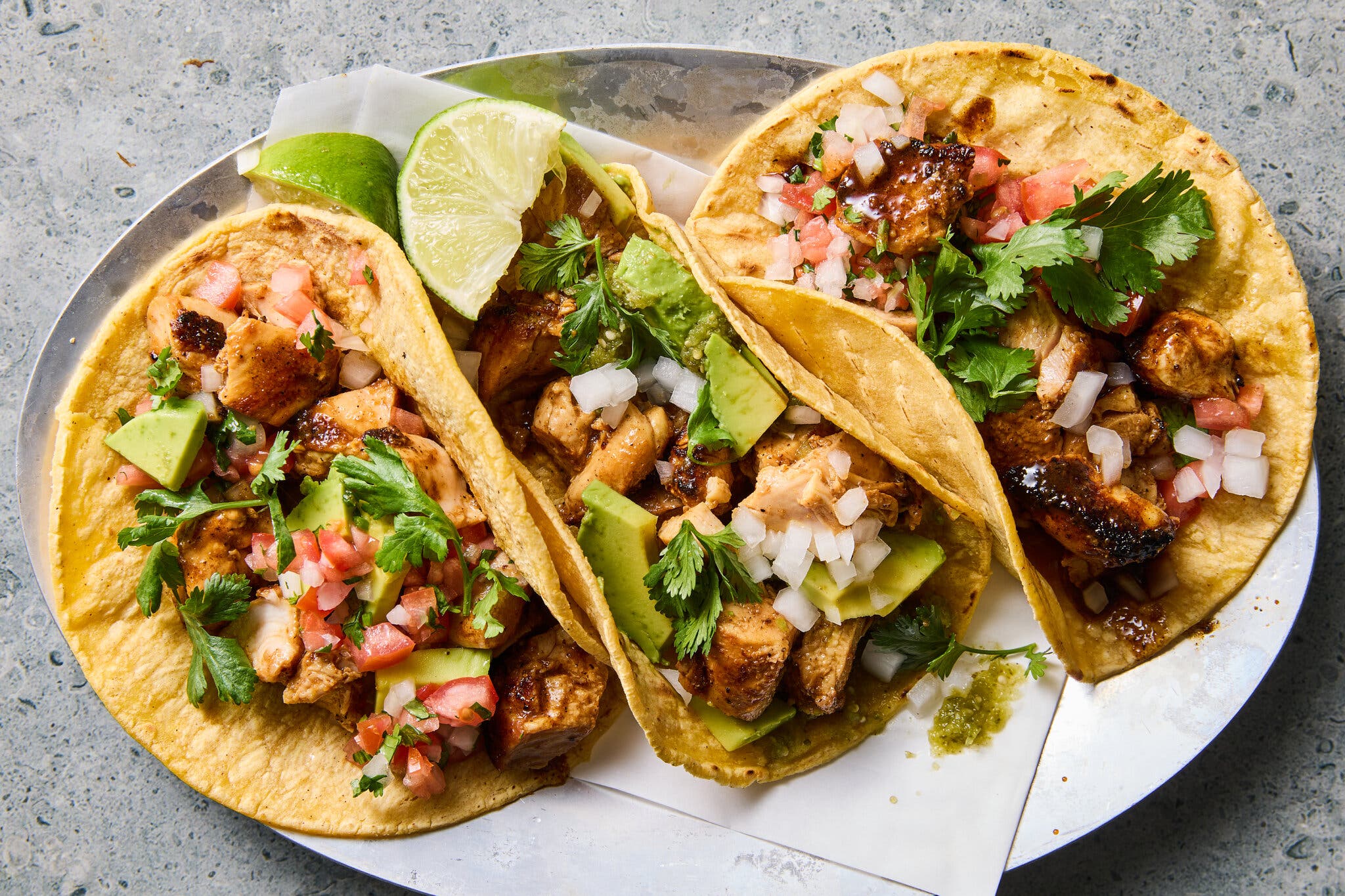 An overhead image of three chicken tacos on a plate topped with herbs, avocado and onions.