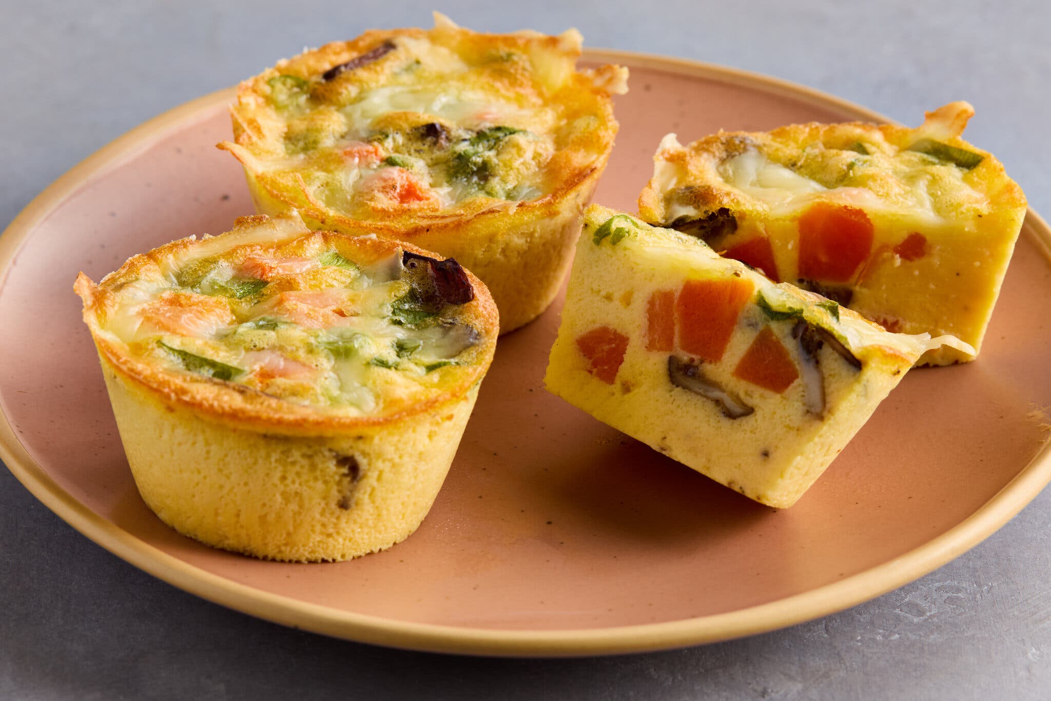 Three baked egg muffins with a golden-brown crust, filled with eggs, chopped vegetables, served on a terracotta-colored plate.