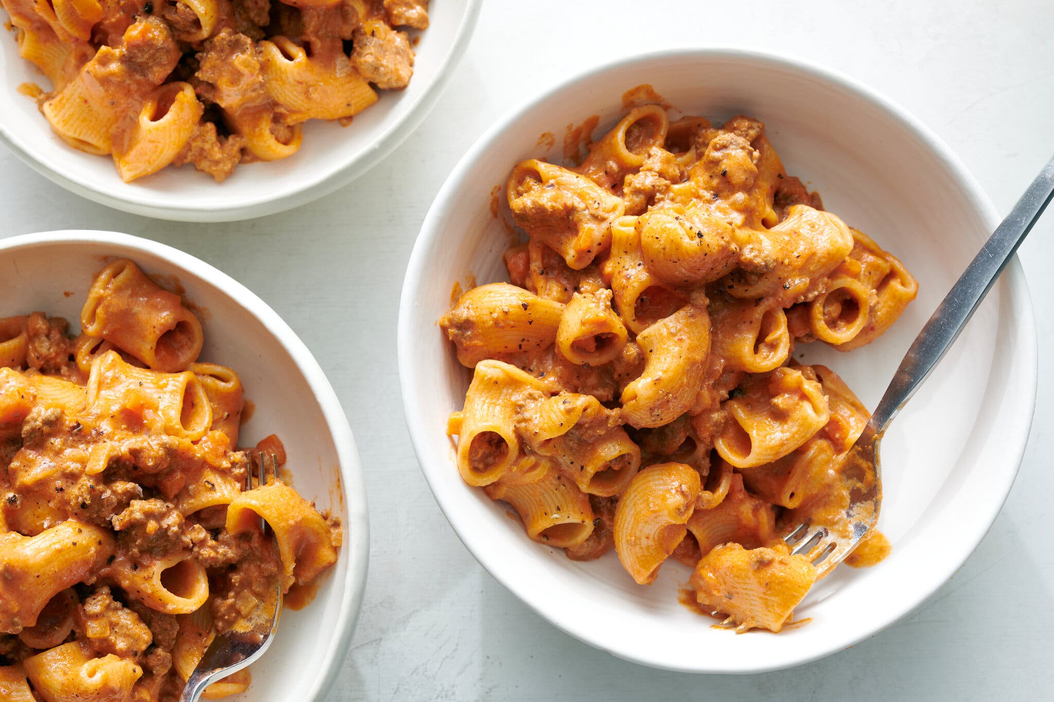 Three white bowls of pasta mixed with a creamy orange sauce and crumbled sausage, with a fork resting in one bowl.