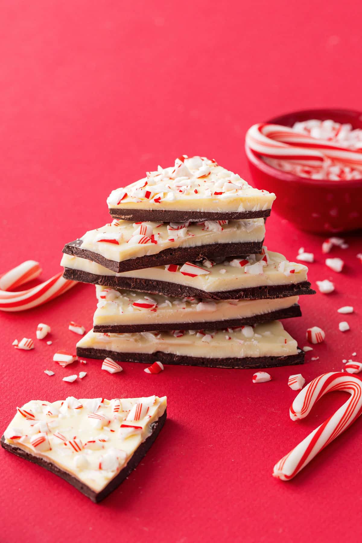 Stack of rough broken pieces of peppermint bark on a red background with candy cane crumbs and a few mini candy canes scattered around.